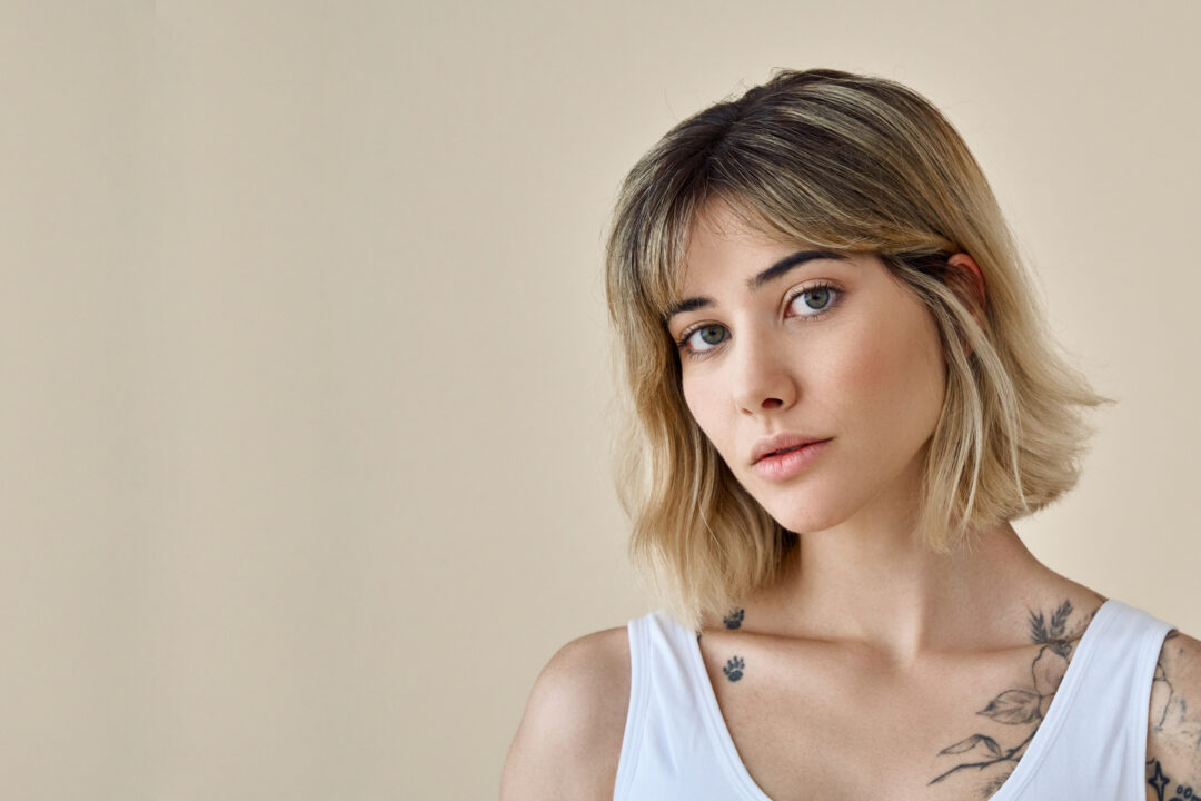 Calligraphy Cut in Düsseldorf, Benrath - Portrait einer blonden jungen Frau mit Tattoos vor einem grauen Hintergrund