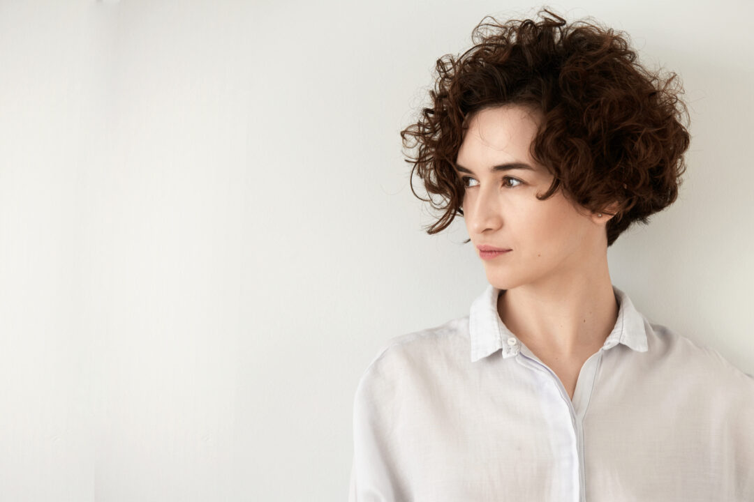 Sascha Mylenbusch - Dauerwelle in Düsseldorf, Benrath - Portrait einer jungen brünetten Frau mit einer lockigen Dauerwelle, vor einem grauen Hintergrund