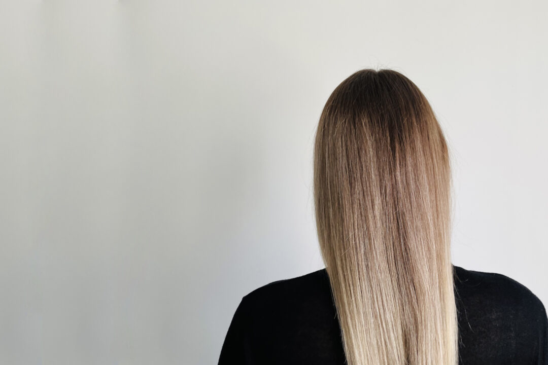 Sascha Mylenbusch Exklusiv Hair & Style - Haarglättung in Düsseldorf, Benrath - Rückenansicht einer blonden jungen Frau vor einem grauen Hintergrund mit gestreckten blonden Haaren