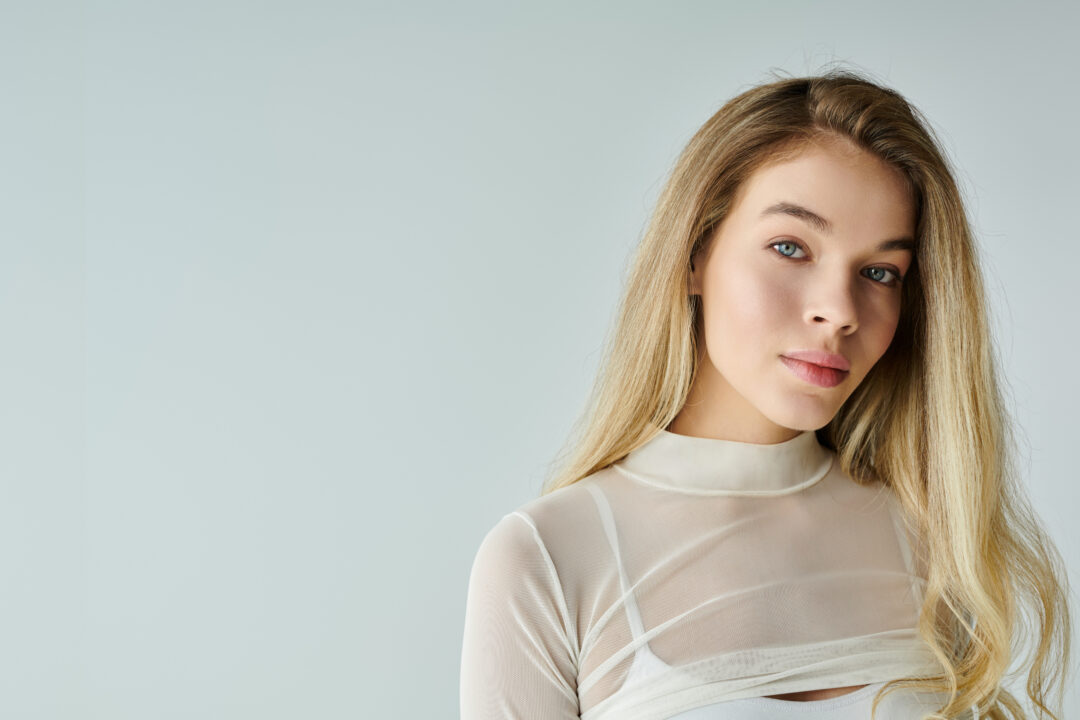 Sascha Mylenbusch Exklusiv Hair & Style - Haarverlängerung in Düsseldorf, Benrath - Portrait einer blonden jungen Frau mit Haarverlängerungen vor einem grauen Hintergrund